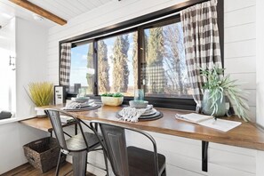 A dining area with a wooden table, two chairs, a window with curtains, and a view of trees. - Tiny Cargo Home, Farm Experience with Rooftop Views (Tulare)