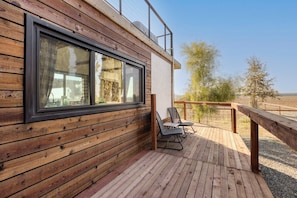 A wooden deck with a view of the countryside, a window with curtains, and two chairs. - Tiny Cargo Home, Farm Experience with Rooftop Views (Tulare)