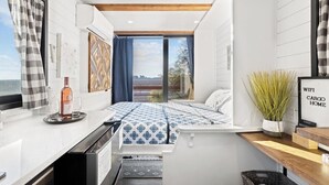 A compact bedroom with a bed, a small kitchenette, a window with curtains, and a view of the outdoors. - Tiny Cargo Home, Farm Experience with Rooftop Views (Tulare)