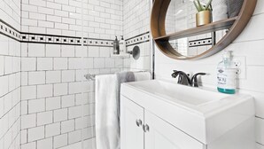 A bathroom with white tiled walls, a white sink with a mirror, and a shower area. - Tiny Cargo Home, Farm Experience with Rooftop Views (Tulare)