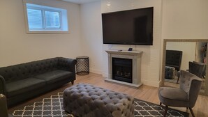 A living room with a dark grey sofa, a grey tufted ottoman, a fireplace, a flat-screen TV mounted above the fireplace, and a large mirror reflecting the room. - Two Bedrooms Modern Cozy Suite (Ardrossan)