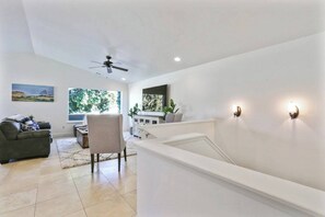 A modern living room with a sofa, a chair, a coffee table, and a dining area with a table and chairs. There is a ceiling fan, recessed lighting, and a large window with a view of greenery. - Slice of Paradise Steps to Morro Rock (Morro Bay)