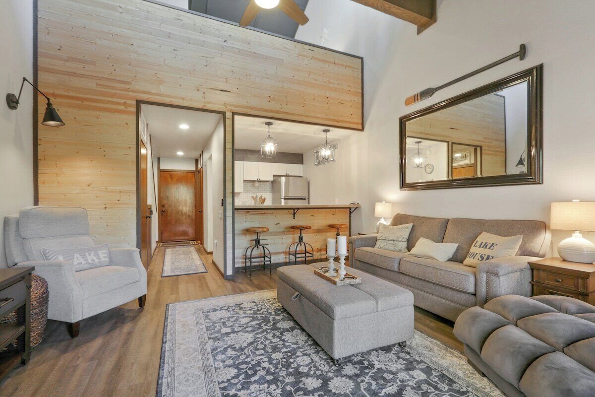 Een moderne woonkamer met een grijze bank, een grijze fauteuil en een getextureerd vloerkleed. Er is een houten plafond en een grote spiegel aan de muur.