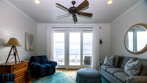 A living room with a ceiling fan, a large window with a view of the ocean, a round mirror, and a blue-striped sofa. - Oceanfront Showplace w/ Private Pool & Elevator; Expansive Beach & Ocean Views; Multiple Porches (Edisto Island)