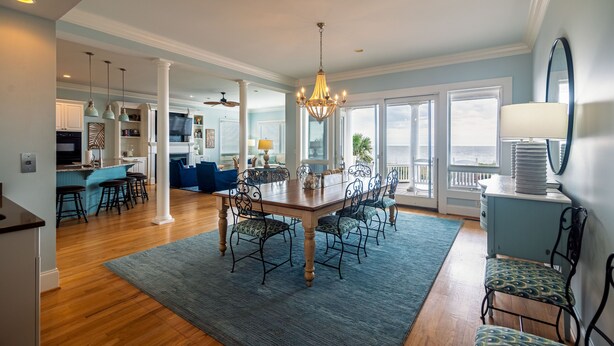 A spacious dining area with a wooden table and chairs, a chandelier, and a view of the ocean through large windows. - Oceanfront Showplace w/ Private Pool & Elevator; Expansive Beach & Ocean Views; Multiple Porches (Edisto Island)