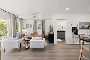 A modern living room with a sofa, two armchairs, a coffee table, and a ceiling fan. There is a kitchen area with a sink, stove, and cabinets visible through an open doorway.