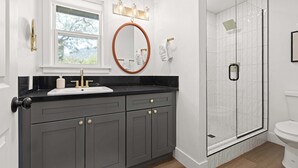 A bathroom with a black countertop, a round mirror, a window, a shower with a glass door, and a toilet. - Riverfront Gateway to Sequoia NP (Three River)