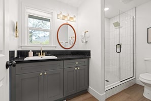 A bathroom with a black countertop, a round mirror, a window, a shower with a glass door, and a toilet.