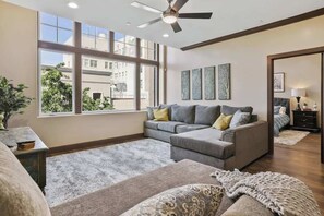 Ein modernes Wohnzimmer mit einem grauen Ecksofa, einem kleinen Tisch und einem großen Fenster mit Blick ins Freie.