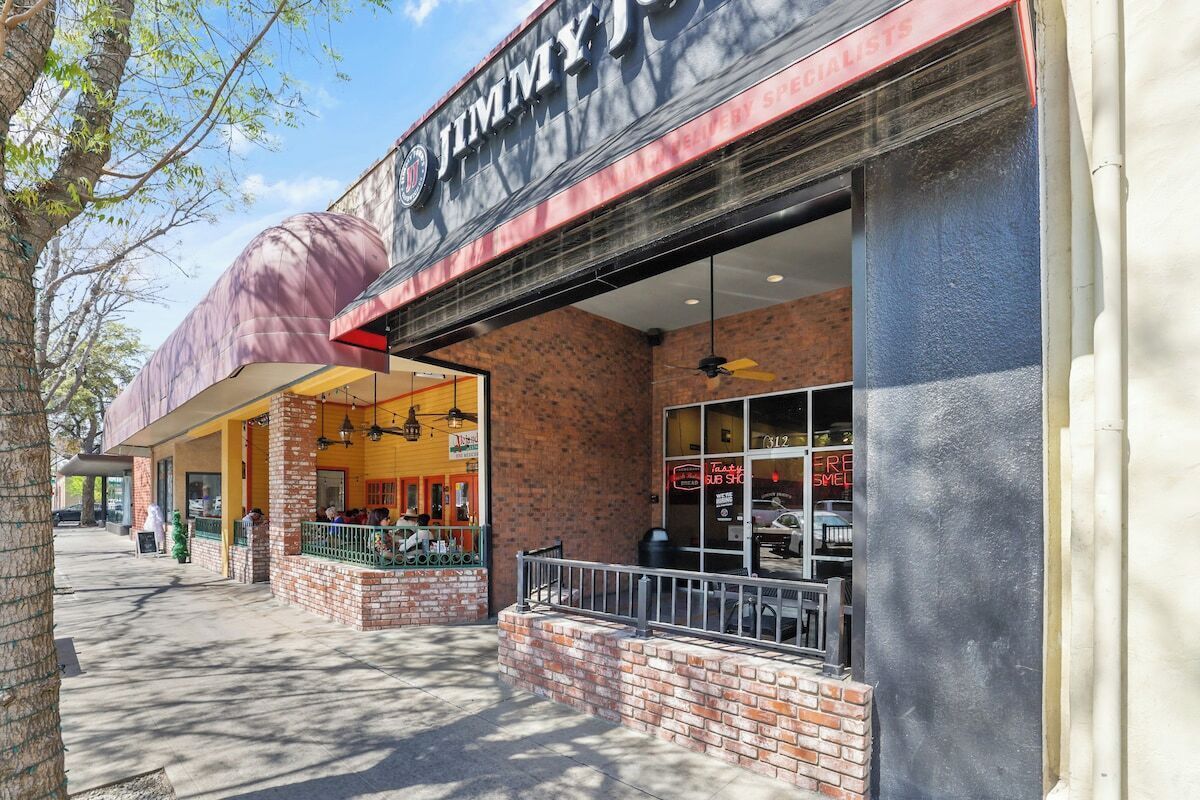 Fachada de Jimmy John’s de ladrillo, con amplios ventanales y mesas al aire libre.