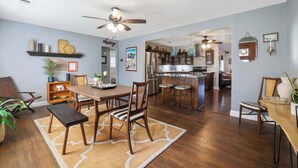 A spacious living area with a dining table, chairs, a ceiling fan, and a kitchen in the background.
