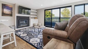 Una sala de estar moderna con chimenea, un sofá de cuero marrón, un tapete estampado y ventanas amplias con vistas al exterior.