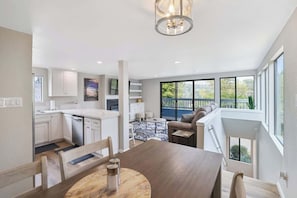 A modern kitchen with white cabinets, a dining area with a wooden table, and a large window with a view of greenery.