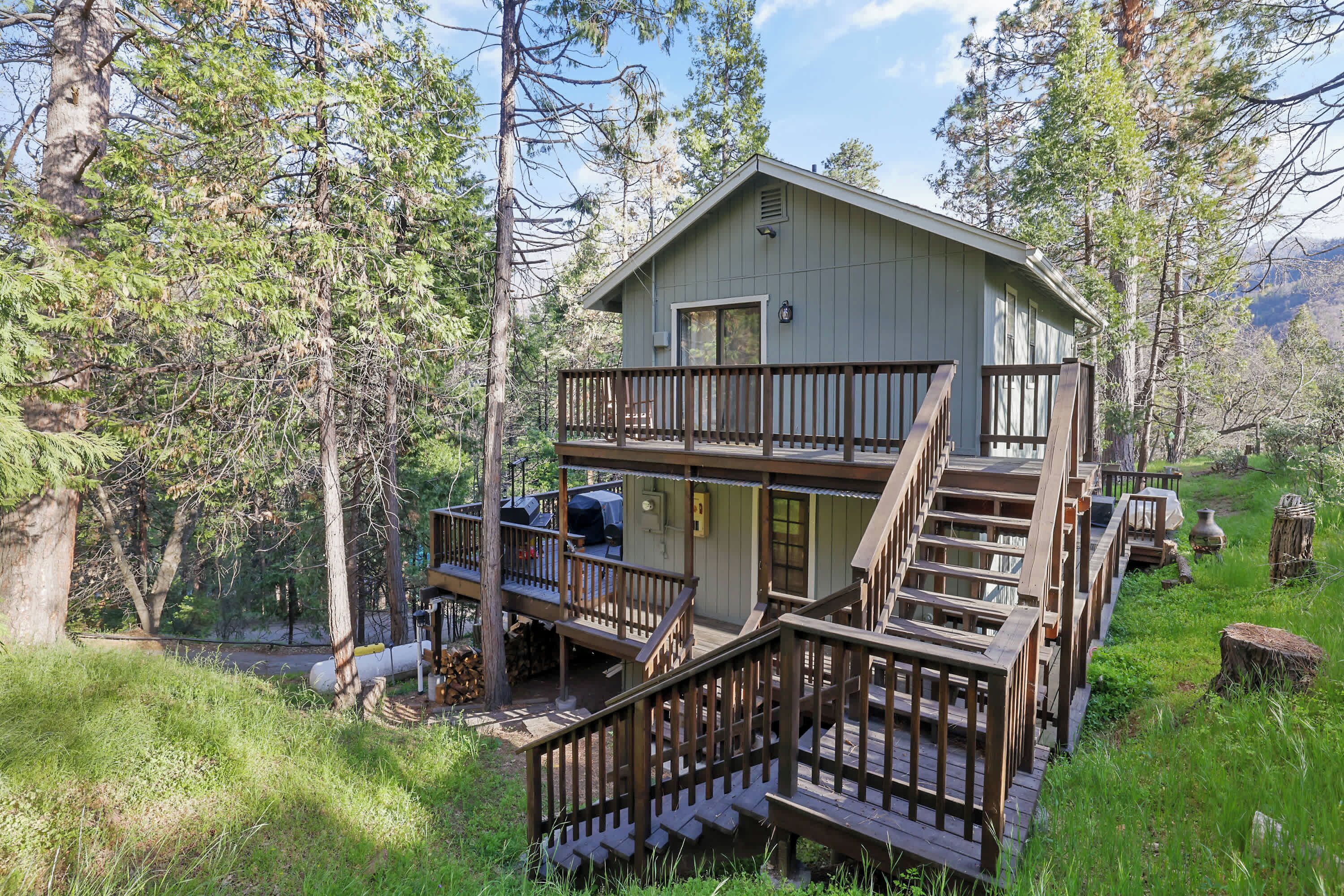 A two-story cabin with a wooden deck and stairs, surrounded by trees.