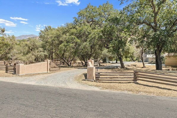 A paved driveway leading to a gated entrance with a metal fence, surrounded by trees and a clear sky.