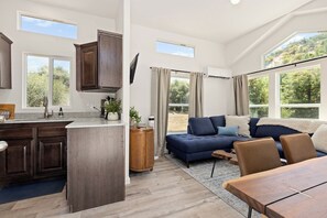 A modern interior with a kitchen, dining area, and living space, featuring large windows and a view of greenery. - Tiny River House w River Frontage (Three River)