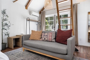 A modern living room with a grey sofa, a small side table, a potted plant, and a window with curtains. - Downtown Retreat w/ Pool & Spa, Near National Parks (Visalia)