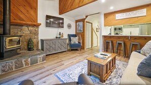 A cozy living room with a stone fireplace, wooden beams, a sofa, and a kitchen area with stools.