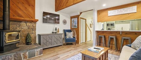 A cozy living room with a stone fireplace, wooden beams, a sofa, and a kitchen area with stools.