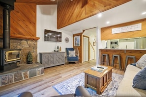 A cozy living room with a stone fireplace, wooden beams, a sofa, and a kitchen area with stools. - Two Bears Basecamp | Views, Trails, Flat Parking (Shaver Lake)
