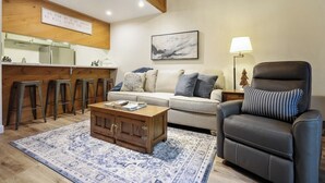 A living room with a sofa, a leather chair, a coffee table, a patterned rug, a kitchen area with stools, and a wall-mounted sign.