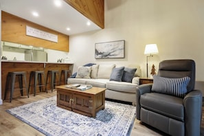 A living room with a sofa, a leather chair, a coffee table, a patterned rug, a kitchen area with stools, and a wall-mounted sign. - Two Bears Basecamp | Views, Trails, Flat Parking (Shaver Lake)