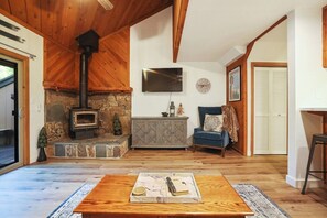 A cozy living room with a stone fireplace, a television, a clock, and a blue armchair. - Two Bears Basecamp | Views, Trails, Flat Parking (Shaver Lake)
