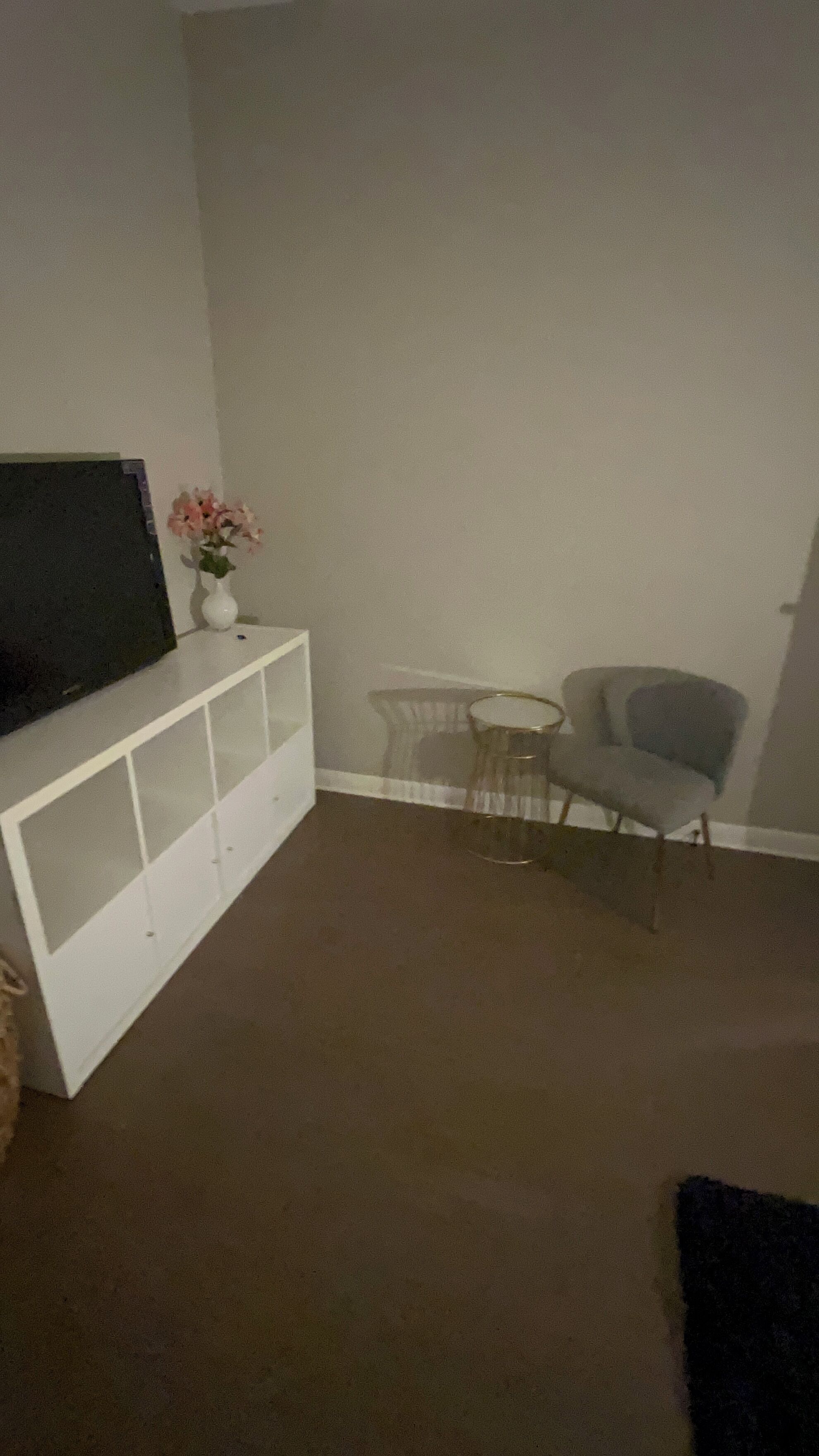 A living room with a flat-screen TV on a white entertainment unit, a small round table with a chair, and a grey armchair.