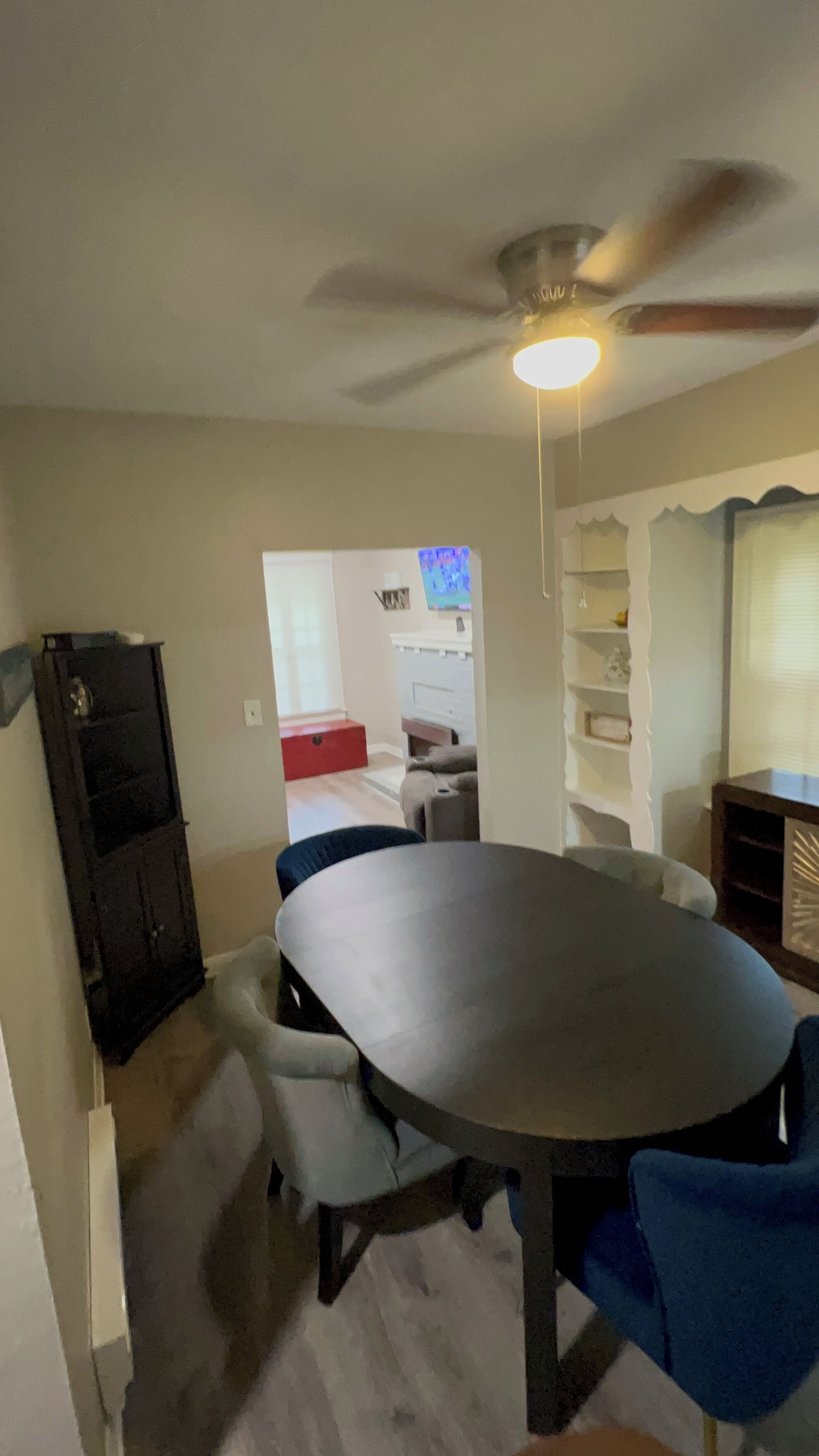 A dining area with a round table, chairs, a ceiling fan, and a view into another room with a TV.