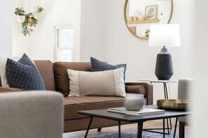 Ein modernes Wohnzimmer mit einem braunen Sofa, gemusterten Kissen, einem kleinen Tisch mit einer Schale, einer Lampe und einer hängenden Pflanze.