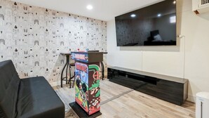 Ein modernes Wohnzimmer mit einer schwarzen Couch, einem farbenfrohen Spielautomaten, einem Flachbildfernseher und Holzboden.