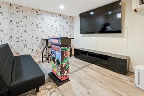 A modern living room with a black sofa, a colorful arcade game cabinet, a flat-screen TV, and a wooden floor.