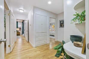 A hallway with wooden flooring, white walls, a white wardrobe with multiple drawers, and a plant on a white shelf.