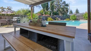 An outdoor patio with a wooden table and bench, a pool, and a backyard with various plants and a barbecue grill. - Visalia Luxe CA: 3BR w/ Private Pool, BBQ, & Patio (Visalia)