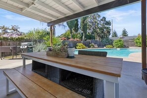 An outdoor patio with a wooden table and bench, a pool, and a backyard with various plants and a barbecue grill. - Visalia Luxe CA: 3BR w/ Private Pool, BBQ, & Patio (Visalia)