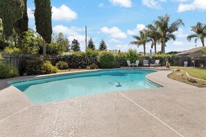 A swimming pool with a surrounding patio area, a wooden fence, and various trees and shrubs.
