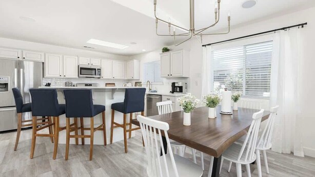 A modern kitchen with white cabinets, a central island with bar stools, a dining table with chairs, and a chandelier.