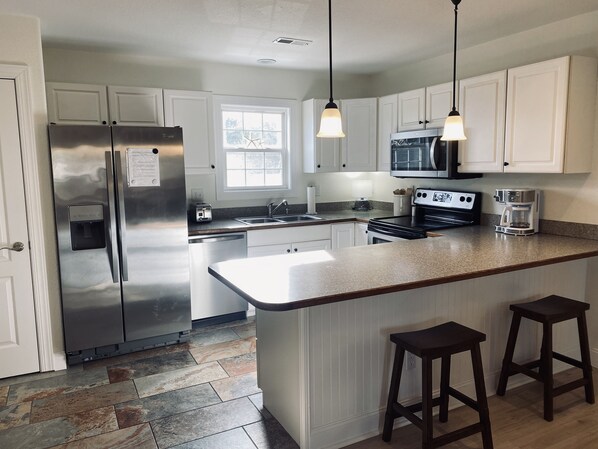 A modern kitchen with white cabinets, stainless steel appliances, a granite countertop, and a large island with stools. - The Grant Atlantic (Kill Devil Hills)