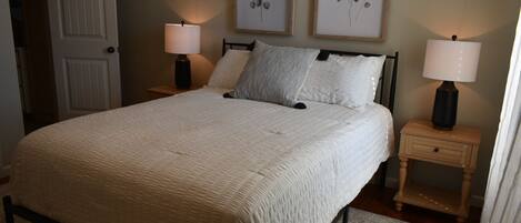 A bedroom with a large bed, two bedside tables with lamps, a door, and two framed botanical prints on the wall.