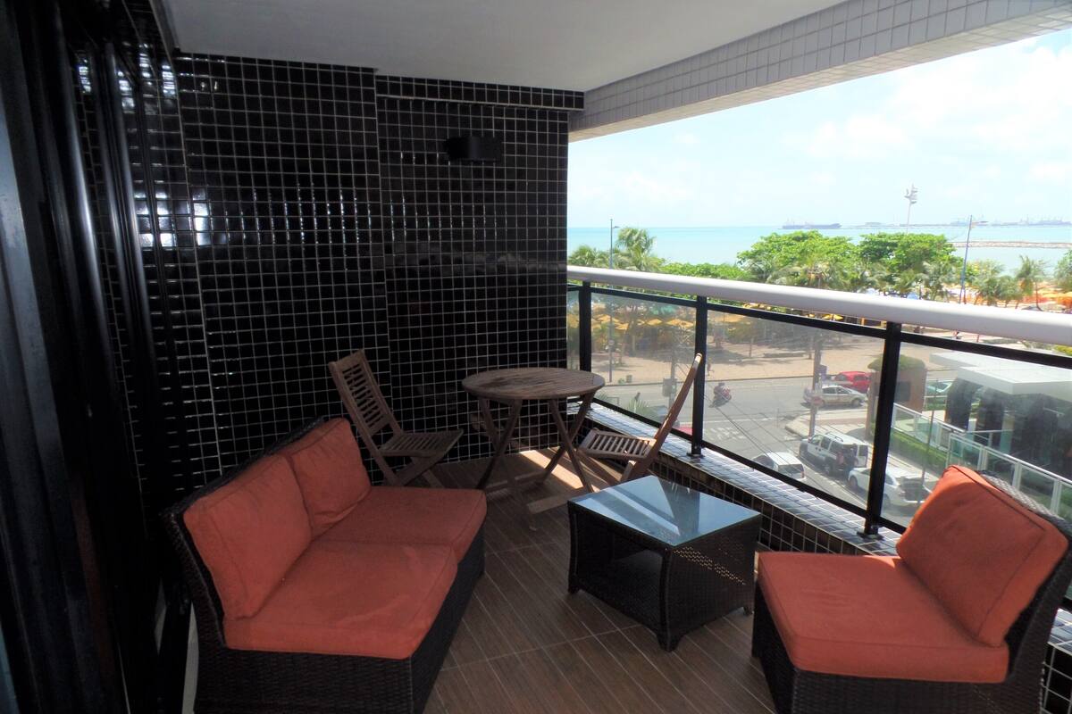A balcony with wicker furniture, a small table, and a view of the sea and cityscape.