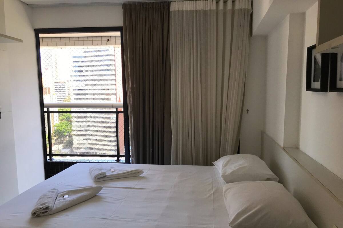 A hotel room with a bed, two pillows, a folded towel, and a window with curtains.