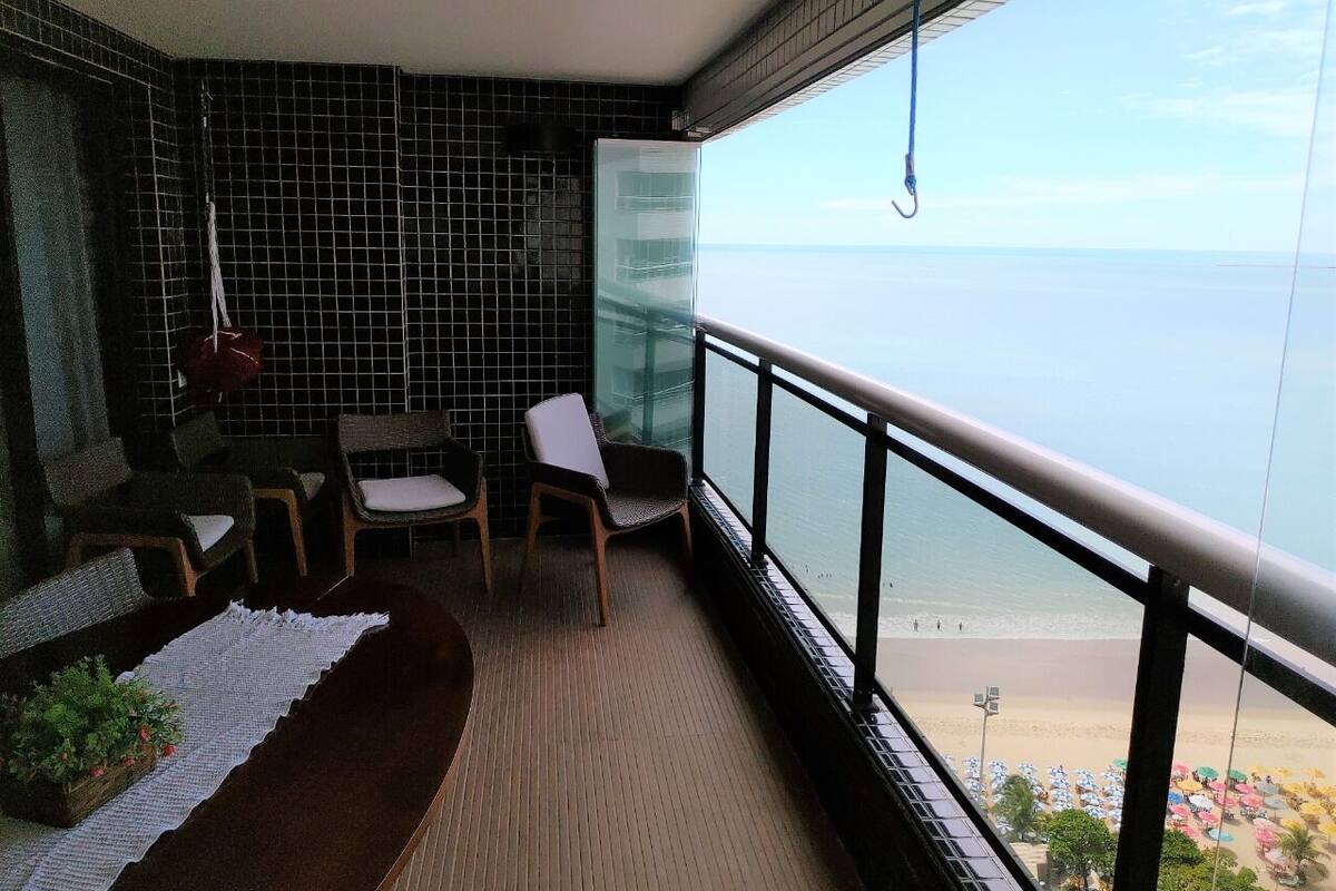 A balcony with a view of the beach, a sofa, two chairs, and a small table with a plant.