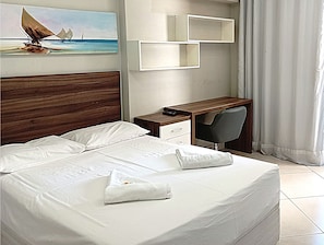 Uma cama bem arrumada com lençóis brancos, cabeceira de madeira, uma escrivaninha com cadeira e um quadro com um veleiro na parede.