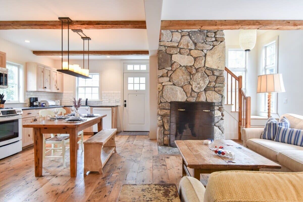 A cozy living room with a stone fireplace, wooden furniture, and a view of the kitchen.