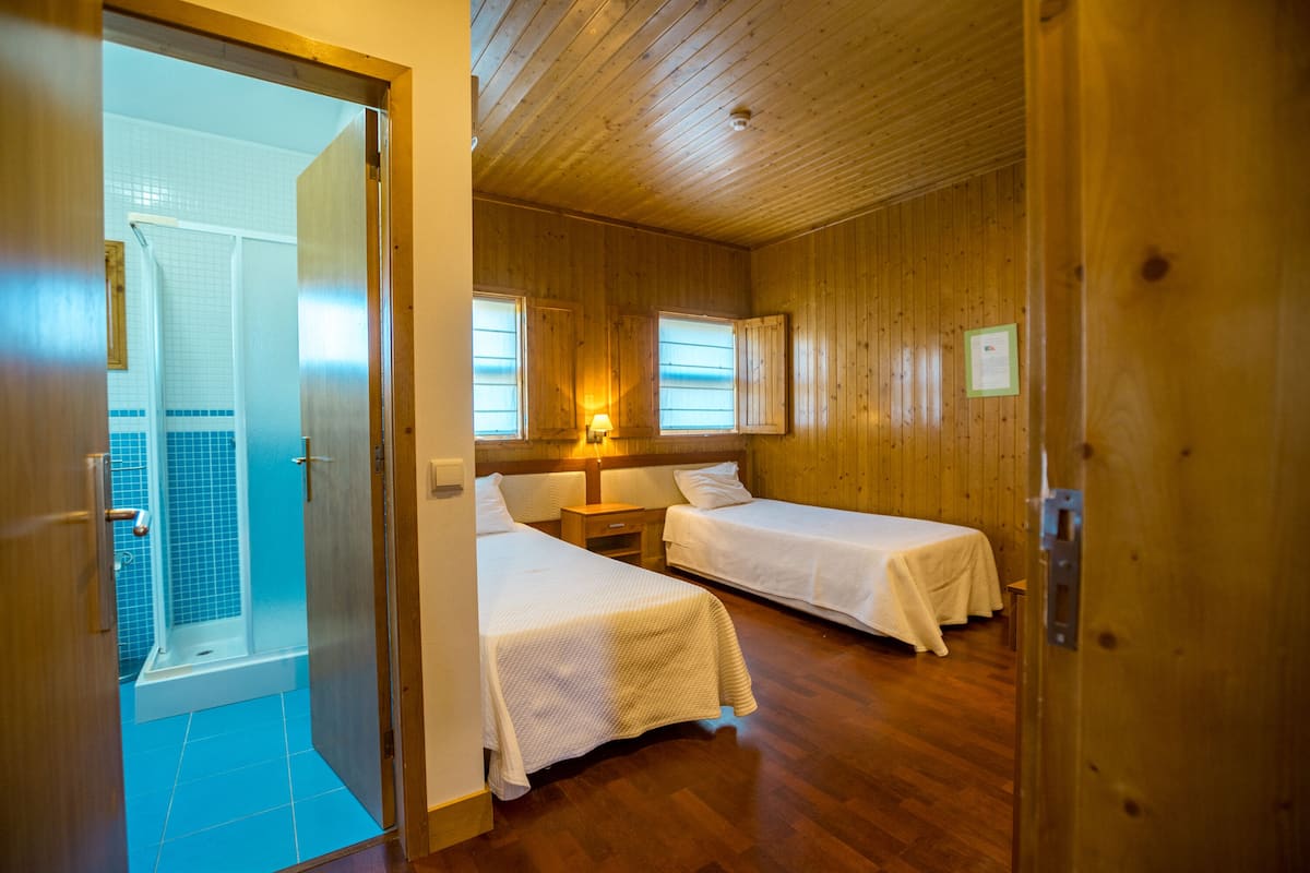 A room with two beds, a wooden ceiling, and a bathroom with blue tiles.