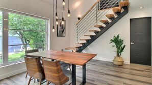 Un interior moderno con una mesa y sillas de madera, una escalera con peldaños de madera y barandales de metal, y vistas al exterior a través de amplios ventanales.