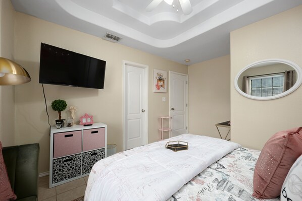 Un dormitorio con cama, televisor montado en la pared, cómoda con cajones, espejo y ventana.