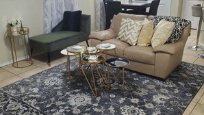 Een woonkamer met een beige bank, een kamerbreed tapijt met dessin, en een eethoek op de achtergrond.