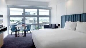 Une chambre d’hôtel moderne dotée d’un grand lit, d’un coin salon avec deux fauteuils, d’une petite table et offrant une vue sur la ville grâce à de grandes fenêtres.