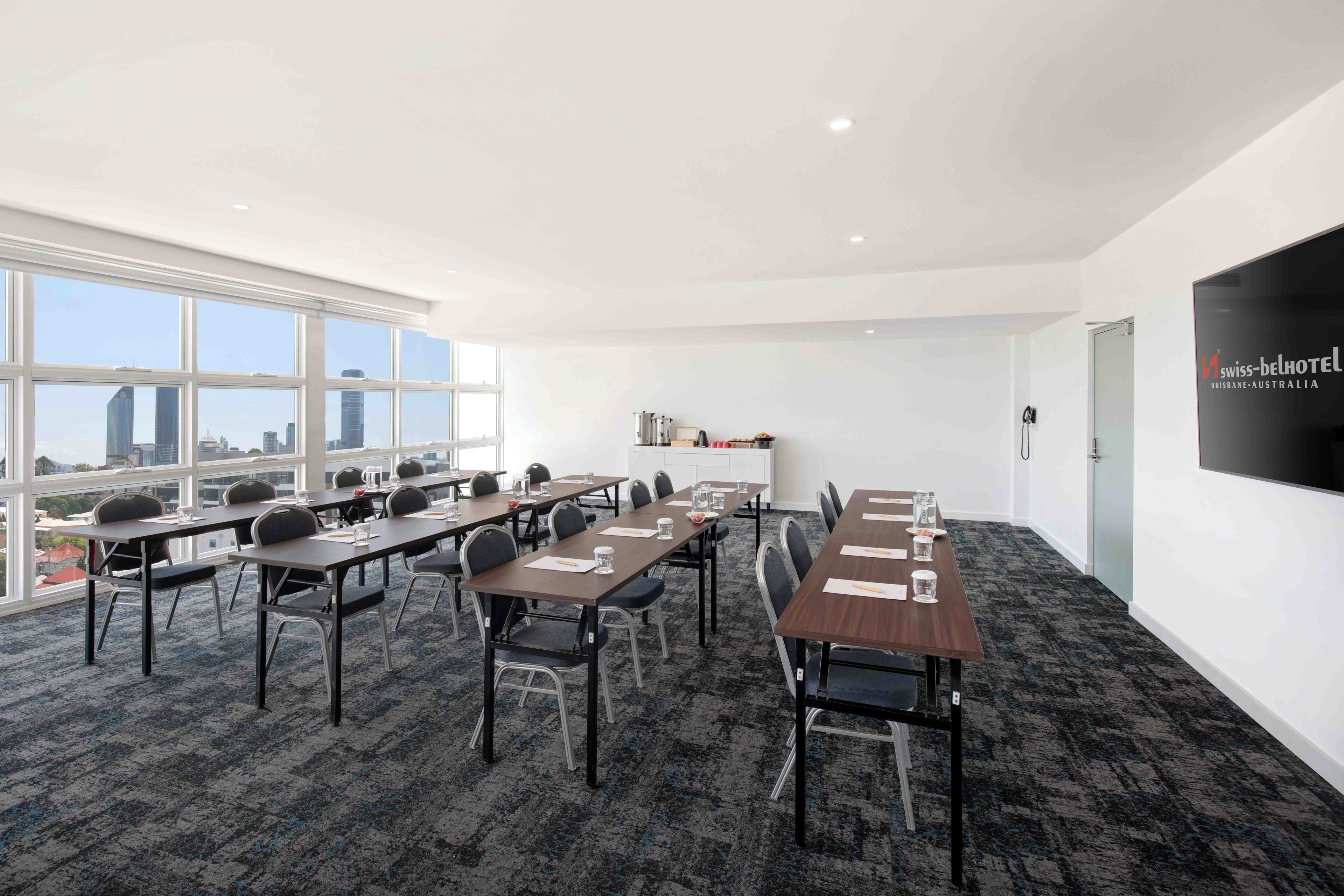A conference room with multiple tables and chairs, large windows with a city view, and a mounted television on the wall.